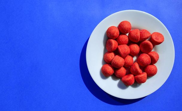 Red strawberry sugary candies in white plate on blue background with copy space - Photo, Image