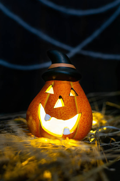 Halloween symbol - glowing Jack o'Lantern pumpkin head with lamp in a pointed black hat in the hay on the background of the cobweb on a dark wooden board, copy space - Photo, Image