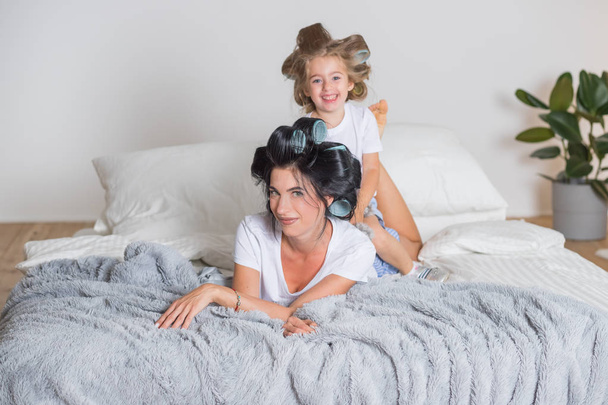 portrét šťastné matky a malé dcery ve vlasech. šťastná máma s dcerou se baví na posteli v pyžamu. Šťastná milující rodina v ložnici ve skandinávském stylu. - Fotografie, Obrázek