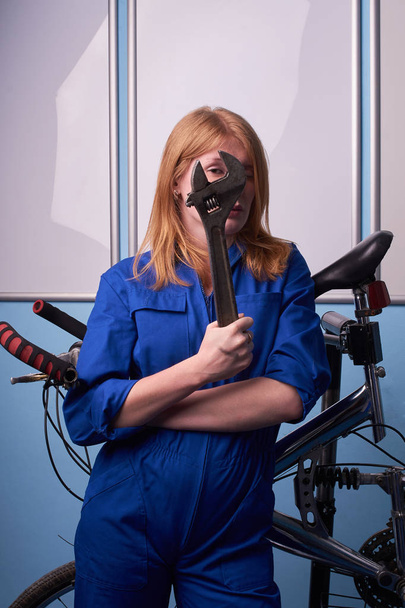Portrait of happy cheerful positive woman who is standing with new bicycle indoors. girl in blue overalls girl with a wrench repairing a bicycle in a bicycle workshop - Foto, immagini