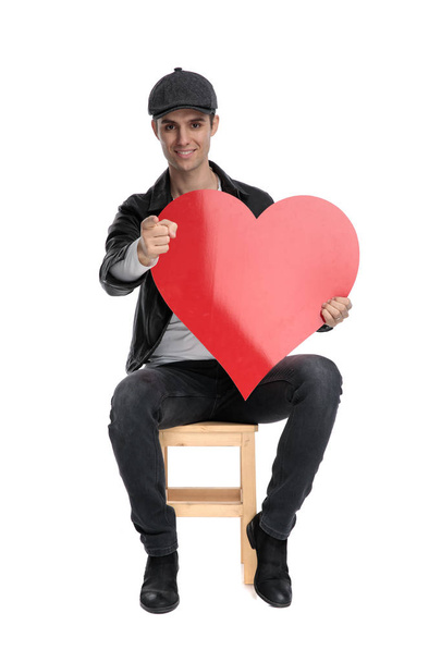 seated casual man holding a big red heart pointing forward - Foto, Imagem