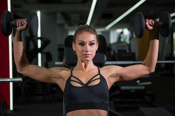 Κορίτσι γυμναστήριο μοντέλο με βαράκια στο γυμναστήριο κάνει ασκήσεις στους ώμους των μυών - Φωτογραφία, εικόνα