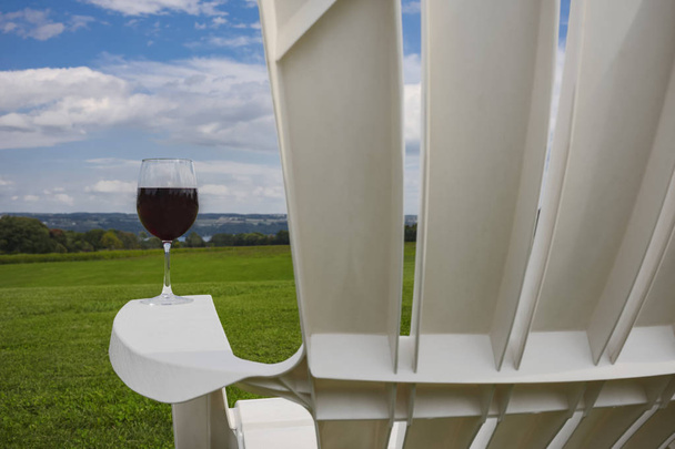 A glass of red wine photographed at on the arm rest of a white, plastic Adirondack chair near Cayuga Lake in the Finger Lakes Region of New York State. - Фото, зображення