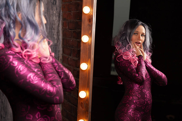όμορφη νεαρή λεπτή γυναίκα στέκεται κοντά σε τεράστιο καθρέφτη με λαμπτήρες - Φωτογραφία, εικόνα