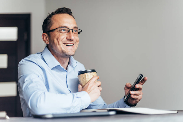 Businessman using phone, holding cup of coffee, sitting at his desk in modern office - Foto, Imagen