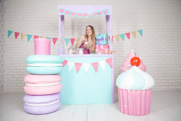 cute happy adult girl posing in fashionable dress among huge fabulous sweets and cakes - Foto, afbeelding