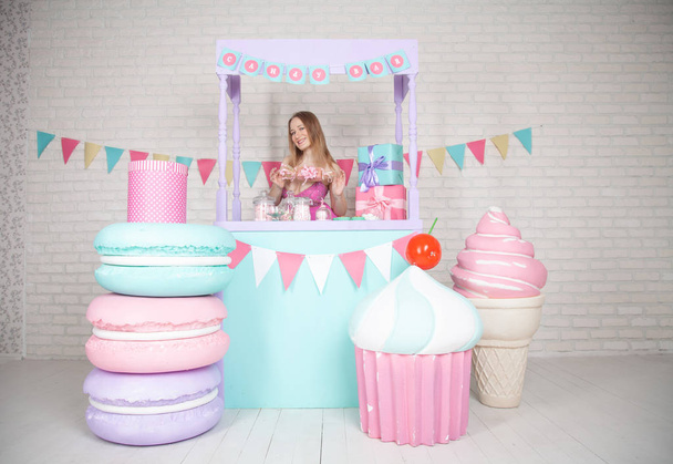 cute happy adult girl posing in fashionable dress among huge fabulous sweets and cakes - Foto, Imagen