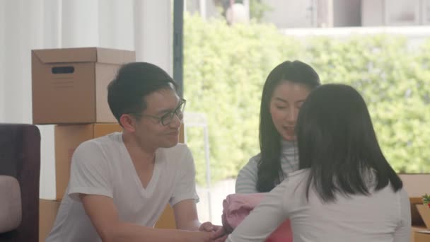 Happy Asian young family relocation removals settle in new home. Excited korean parents unpack cardboard boxes together with little daughter holding clothing in living room at house on moving day. - Metraje, vídeo