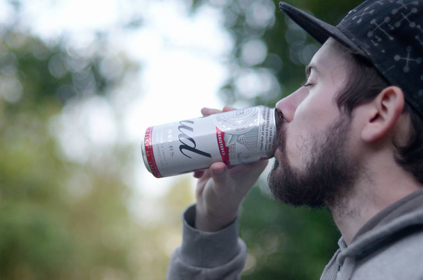 Joven barbudo bebe cerveza Budweiser Bud en lata de aluminio sobre árboles verdes fondo borroso
 - Foto, Imagen