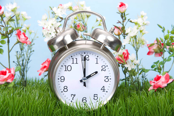 Retro style clock sitting in green grass with flowers representing daylight savings spring forwards. - Photo, Image