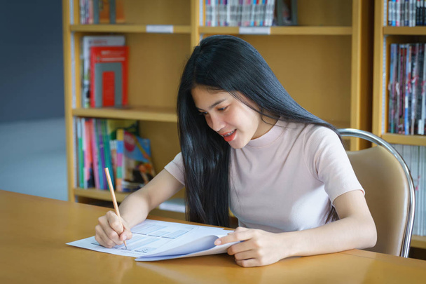 Νεαρή φοιτήτρια Πανεπιστημίου επικεντρώνεται στην εξέταση της γλωσσικής πρακτικής μέσα στη βιβλιοθήκη. Κορίτσι φοιτητής γράφει την άσκηση των εξετάσεων - Φωτογραφία, εικόνα