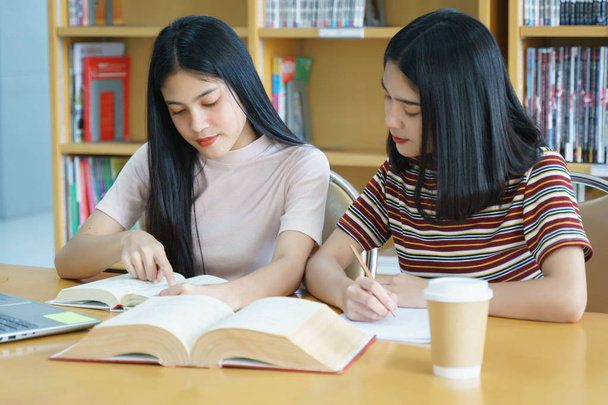 Młody Asian kobieta studia studenckie i bierze notatki z książki w bibliotece. Studentka Uniwersytetu siedzącego przy stole robi zadania w bibliotece kolegium. Student za pomocą laptopa i uczenia się online. - Zdjęcie, obraz