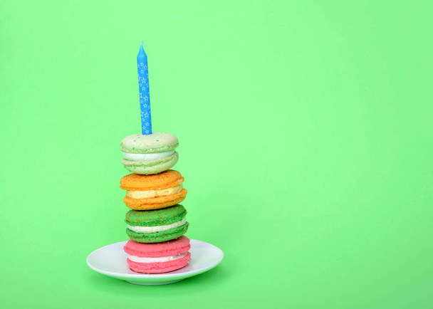 pilha de biscoitos macaron com creme de enchimento em uma placa branca com vela azul no fundo verde. Festa de aniversário tema
. - Foto, Imagem