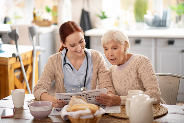 Caregiver reading morning news for retired woman - Foto, Imagen