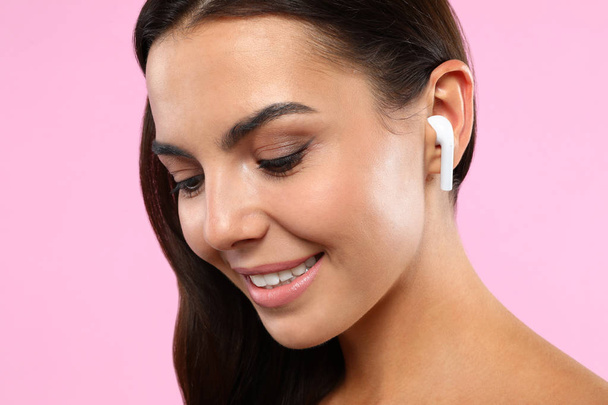 Happy young woman listening to music through wireless earphones on pink background, closeup - Foto, afbeelding