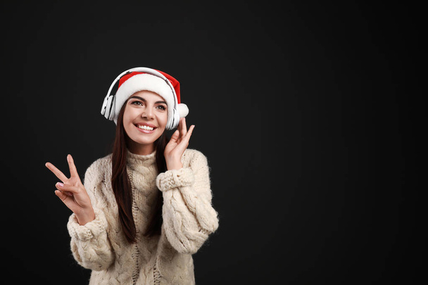 Young woman in Santa hat listening to Christmas music on black background, space for text - Zdjęcie, obraz