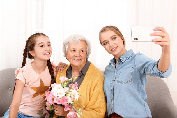 Sœurs heureuses prenant selfie avec leur grand-mère tenant des fleurs à la maison
 - Photo, image