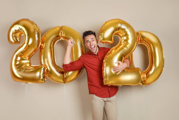 Excited young man with golden 2020 balloons on beige background. New Year celebration - Zdjęcie, obraz