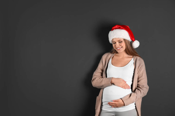 Young pregnant woman in Santa hat on grey background, space for text. Christmas celebration - Foto, Bild