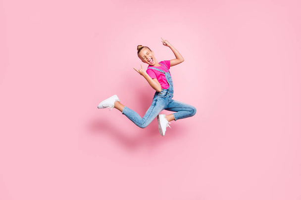 Full length body size photo stylish trendy cute cheerful rejoicing girl wearing jeans denim overall t-shirt fuchsia jumping showing rock sign isolated over pink pastel color background - Foto, imagen