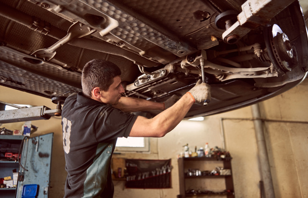 Mécanicien de voiture spécialiste expérimenté debout sous la voiture relevée pendant le processus de réparation et d'entretien dans le garage
 - Photo, image