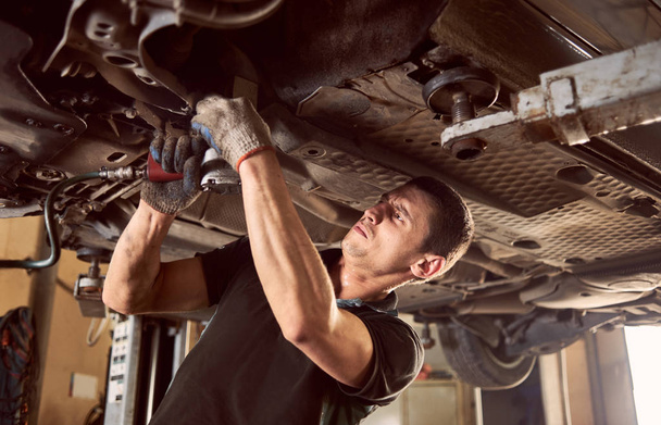 Ervaren specialist automonteur staat onder opgeheven auto tijdens reparatie- en onderhoudsproces in reparatiepost - Foto, afbeelding