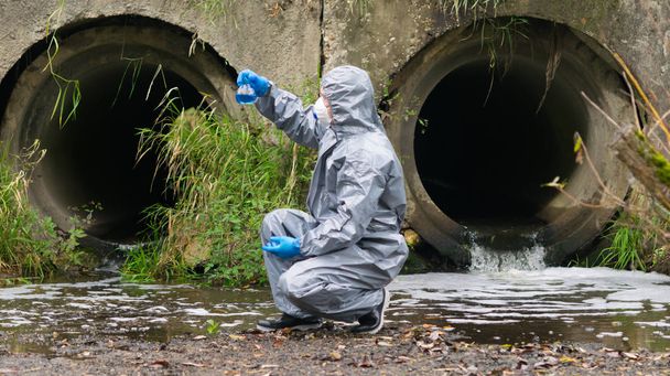 a man in protective clothing, looking at a tube of blue liquid, against the drainpipes - Fotó, kép