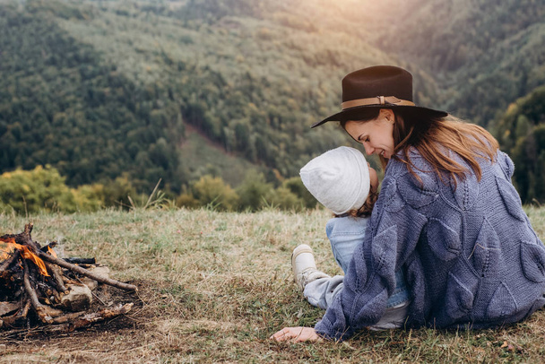 Eine junge Mutter und ihre hübsche Tochter sitzen am Lagerfeuer in den Bergen im wilden Nationalpark-Wald. Reisende Familie erholt sich nach dem Bergsteigen. Kaukasische Hündin mit Baby in den Bergen - Foto, Bild