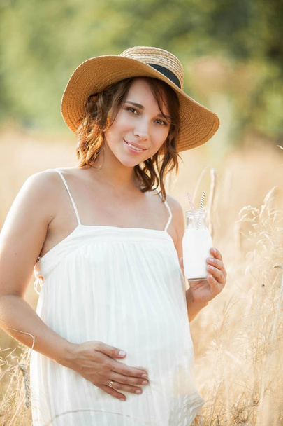 Beautiful young pregnant woman in a wheat field drinks milk from a glass bottle. Portrait of a young pregnant woman in the sun. Summer. - 写真・画像