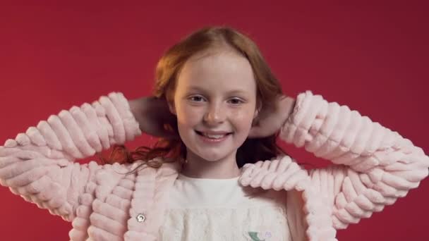 Close up of adorable happy girl playing with her red curly hair and looking at camera on the red wall - Filmagem, Vídeo