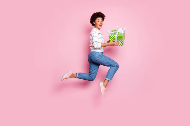 Full length body size photo of excited fast quick crazy funny girlfriend black skinned cute cheerful wearing jeans denim striped sweater holding giftbox with hands isolated pastel color background - Φωτογραφία, εικόνα