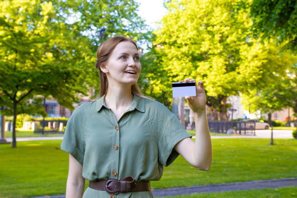 Молодая блондинка в зеленом платье с кредиткой на фоне парка
 - Фото, изображение