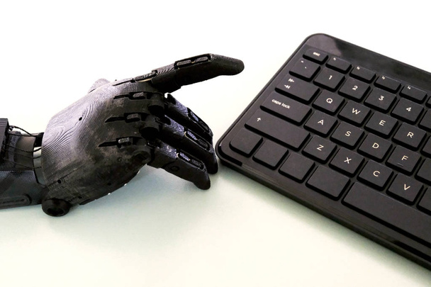 Zwarte robot wijzers en computer toetsenbord op een lichte tafel achtergrond. De prothese van een menselijke hand verhoogde zijn wijsvinger om op een toets te drukken. Het concept van digitalisering en cybernetica. - Foto, afbeelding