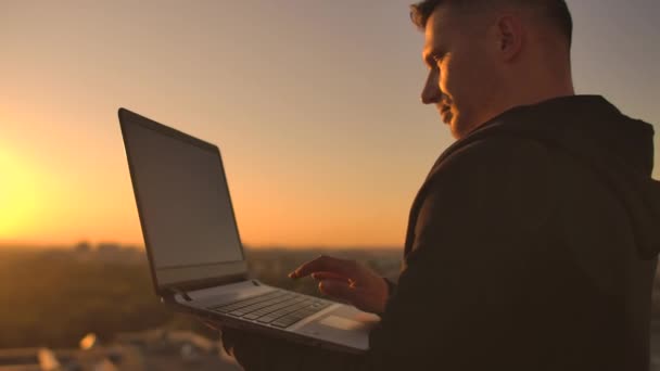 Close-up: um programador digitando a mão em um teclado de laptop ao pôr do sol com vista para o telhado. Um homem de negócios trabalha remotamente. Freelancer executa trabalho em férias
. - Filmagem, Vídeo