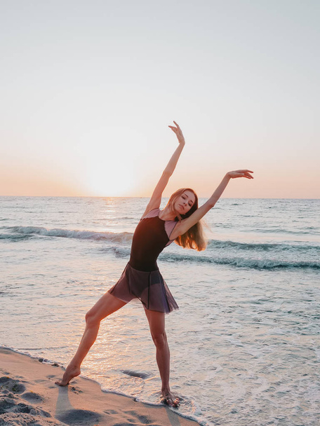 Flexible cute ballerina in black dress dancing ballet on sea sandy beach in morning light. Concept of stretching, art, nature beauty - Foto, immagini