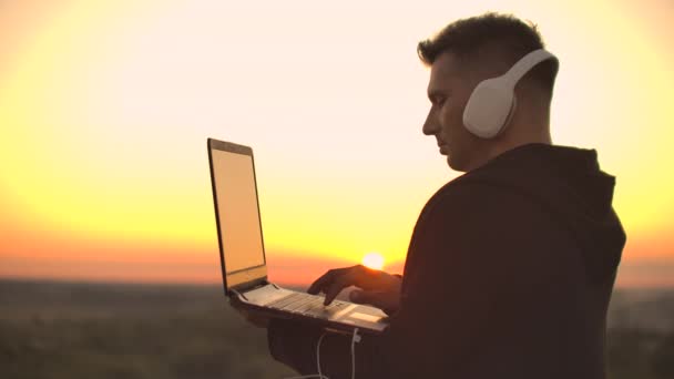 A man in headphones on the roof relaxes working remotely enjoying life despite a handsome kind of sipping beer and types on the keyboard. - Footage, Video