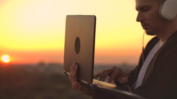 Чоловік-фрілансер у навушниках, що стоять на даху під час заходу сонця, пише на сторінках коду клавіатури. Маленький бізнес. Слухайте музику і працюйте за комп'ютером, насолоджуючись прекрасним видом з даху
 - Кадри, відео