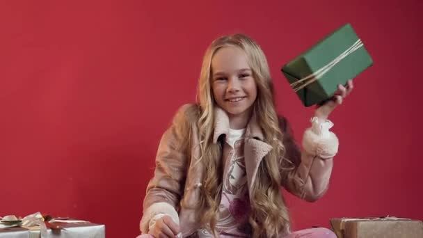 Slow motion of adorable smiling teen girl in stylish clothes holding in her hand green box with gift and looking at camera on the red background - Záběry, video