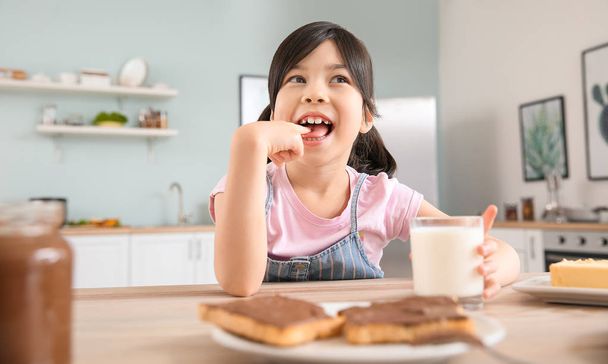 Забавная маленькая девочка ест вкусные тосты с шоколадом, распространяющимся на кухне
 - Фото, изображение