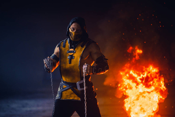 Человек, изображающий воина Скорпиона в цепях, позирует перед огнём
 - Фото, изображение