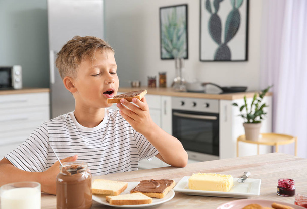 Funny little boy eating tasty toasts with chocolate spreading in kitchen - Photo, Image