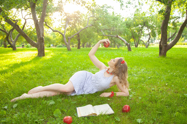 Valkoihoinen onnellinen opiskelija tyttö söpö valkoinen mekko levätä vihreä kesä ruoho kirja ja punainen omenat
 - Valokuva, kuva