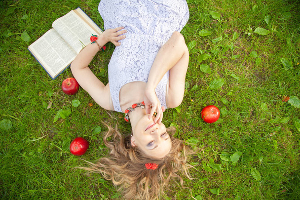 καυκάσιος χαρούμενος φοιτητής κορίτσι σε χαριτωμένο λευκό φόρεμα υπόλοιπο στο πράσινο γρασίδι του καλοκαιριού με το βιβλίο και κόκκινα μήλα - Φωτογραφία, εικόνα