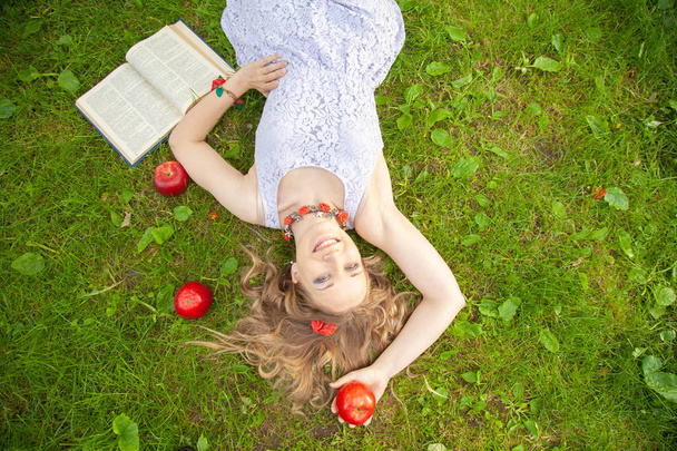 kaukasische glückliche Studentin in niedlichem weißen Kleid ruhen auf dem grünen Sommergras mit Buch und roten Äpfeln - Foto, Bild