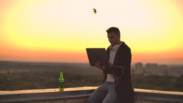ein Mann mit Kopfhörern auf dem Dach entspannt sich bei der Arbeit und genießt das Leben, trotz einer hübschen Art Bier zu schlürfen und auf der Tastatur zu tippen. - Filmmaterial, Video
