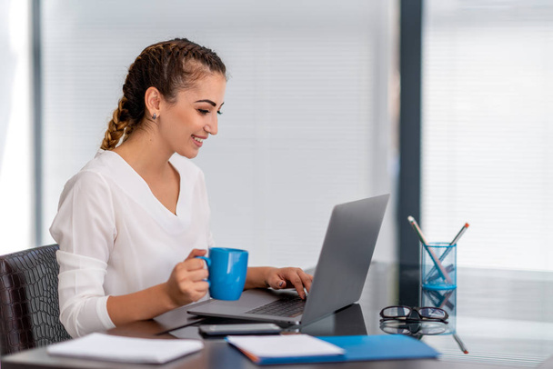  Cheerfull young business woman sitting at her desk and drinking tea - Foto, Imagen