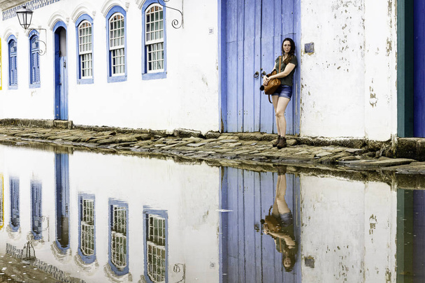 Hermosa modelo de pie en la puerta de madera azul en la histórica ciudad de Paraty, Río de Janeiro, Brasil
. - Foto, Imagen