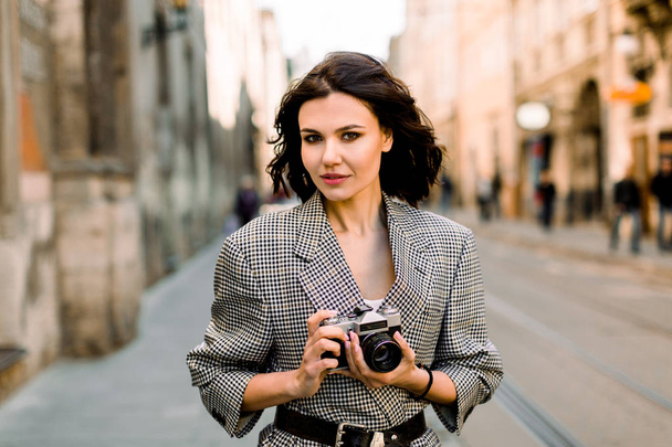 Portrait of pretty brunette girl with short hair holding retro camera and posing on the street in city. She wears gray checkered acket, and has nude makeup. - Foto, Bild