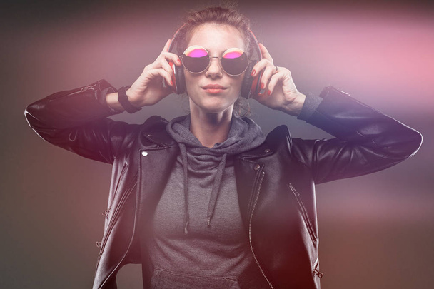 divatos fiatal nő piros fejhallgató pózol bőr kabát és napszemüveg ellen sötét háttér - Fotó, kép