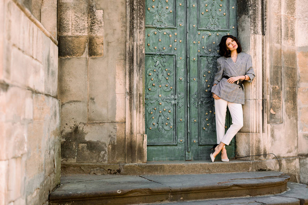 fashion shot of pretty young woman near old ancient door, she is looking at camera, wearing stylish jacket and white pants - Foto, Bild